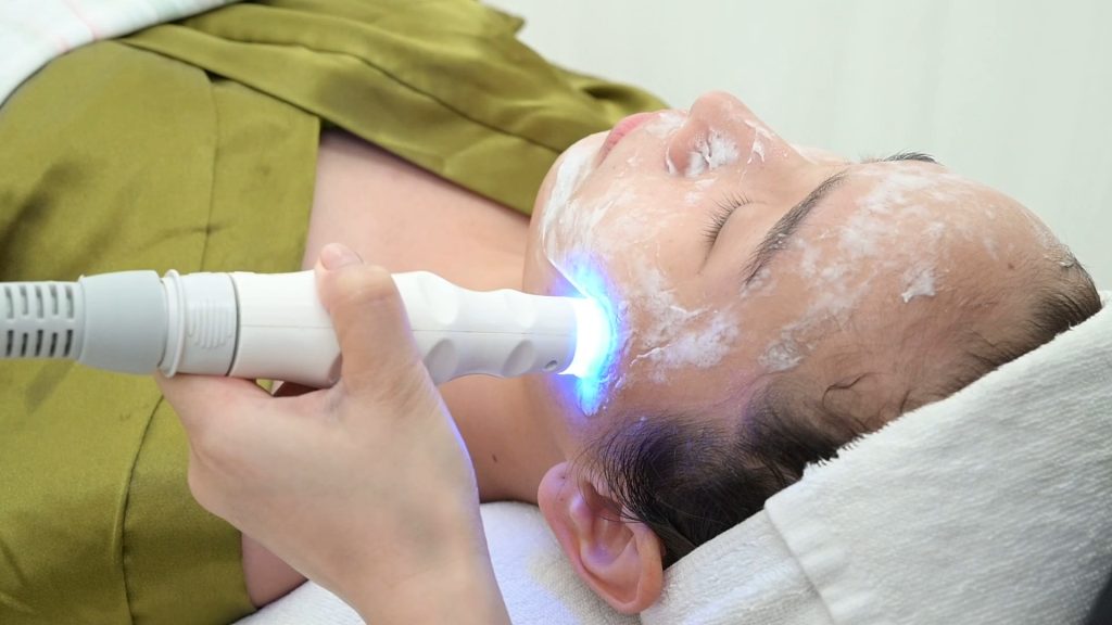 Đầu máy phát ánh sáng xanh được đưa lên khuôn mặt khách đang phủ lớp gel trong — Lona Home Spa, Cần Thơ