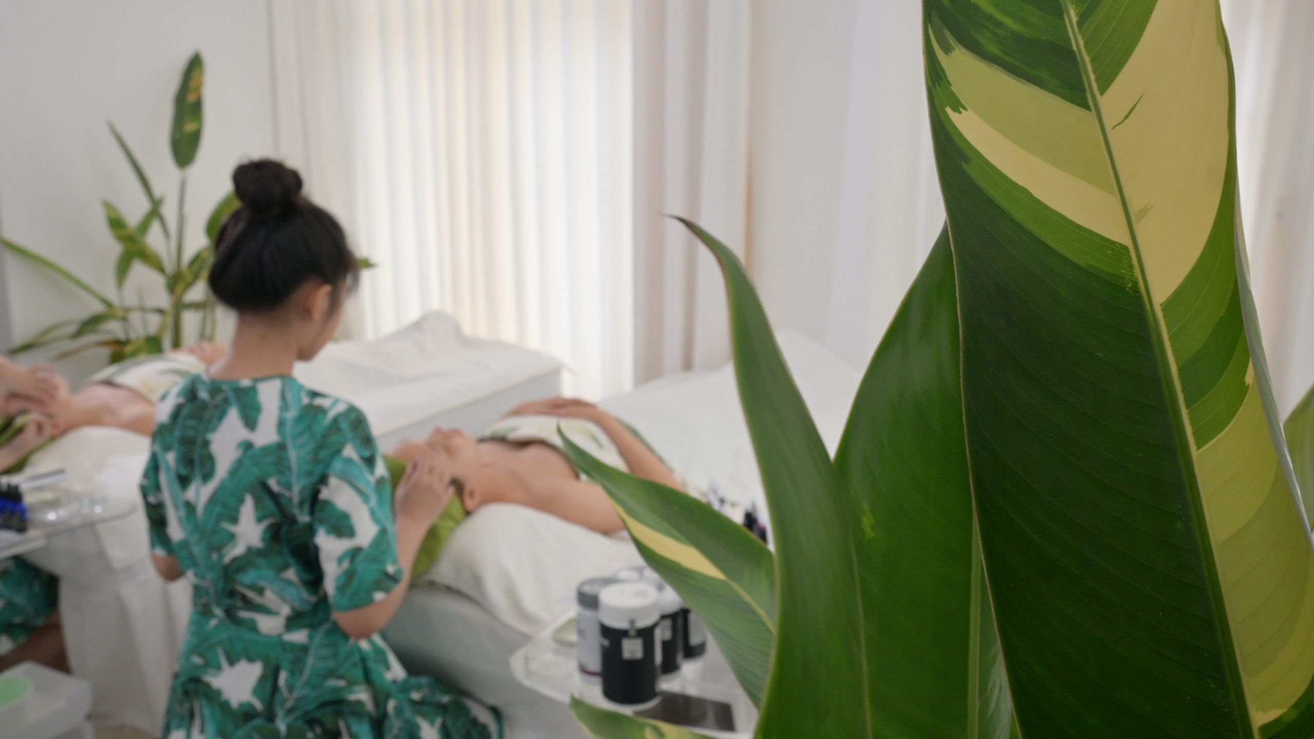 Kỹ thuật viên mặc đồng phục hoạ tiết lá chuối chuẩn bị giường trị liệu, bên cạnh là cây xanh — Lona Home Spa, Cần Thơ